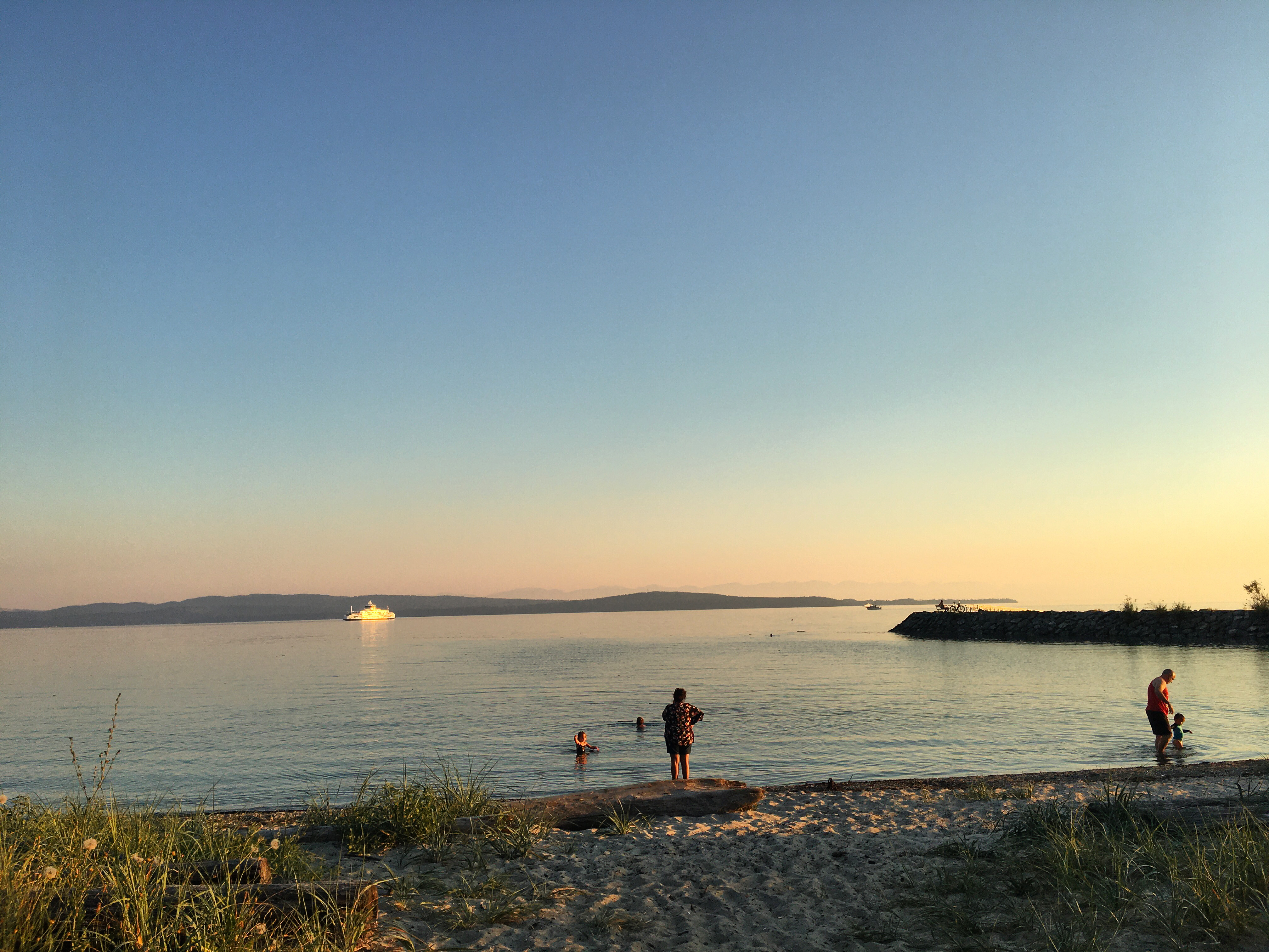 A beach in Powell River, BC at sunset. There's a ferry moving in front of Texada Island. A breakwater juts out from the right of the frame. People are swimming in the ocena.